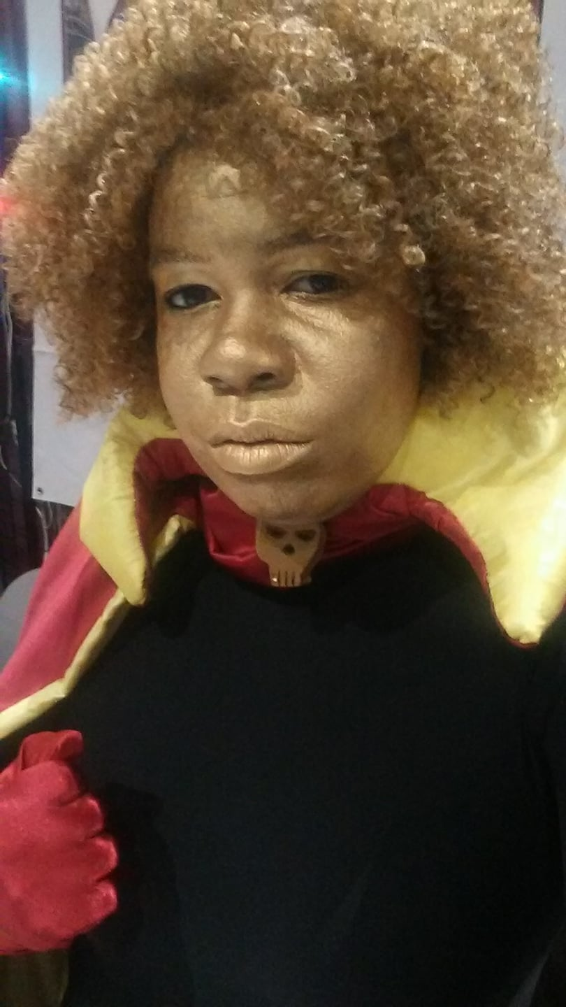 close up of Jay as Adam Warlock, with gold skin, curly blond hair, a black body suit under a red cape with yellow lining, a skull broach, and red gloves