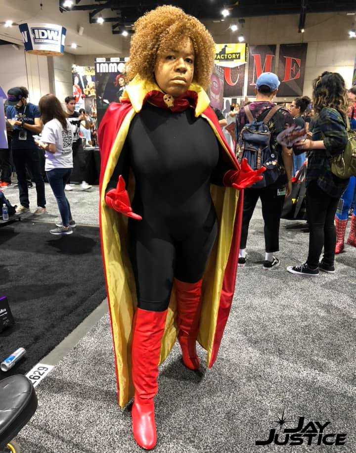full length photo of Jay as Adam Warlock, with gold skin, curly blond hair, a black body suit under a red cape with yellow lining, a skull broach, red gloves and red boots